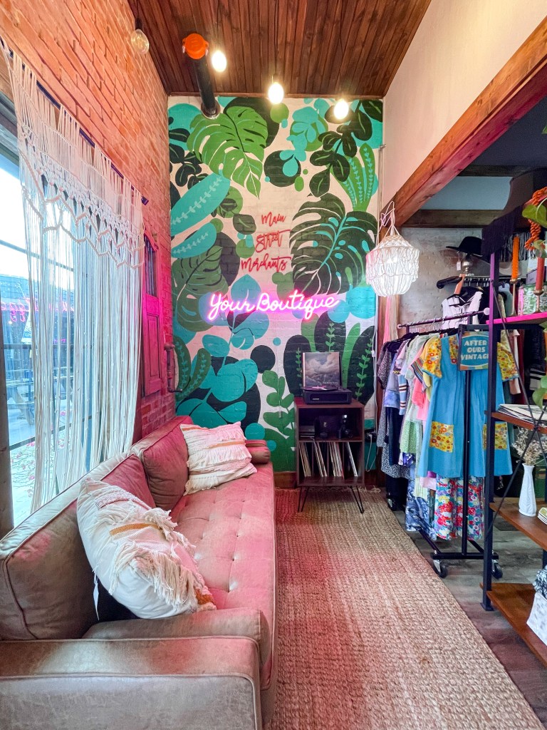 A seating area at a woman's clothing boutique with a mural of plants with a neon sign reading "your boutiqur"