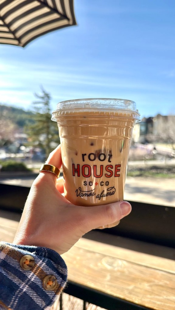 Woman holding up an iced coffee cup from Root house coffee in pagosa springs, Colorado with a view of the river in the background